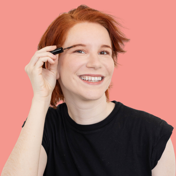 Redhead holding a Redhead Revolution Eyebrow Gel wand against a pink
 background