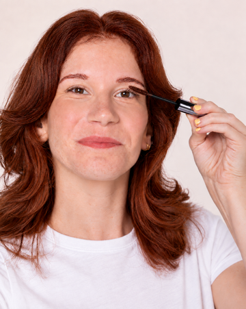 A woman with shoulder-length auburn hair applies Redhead Revolution's Gingerlash Mascara to her eyelashes, holding the black applicator wand. She is wearing a white t-shirt against a plain, light beige background.