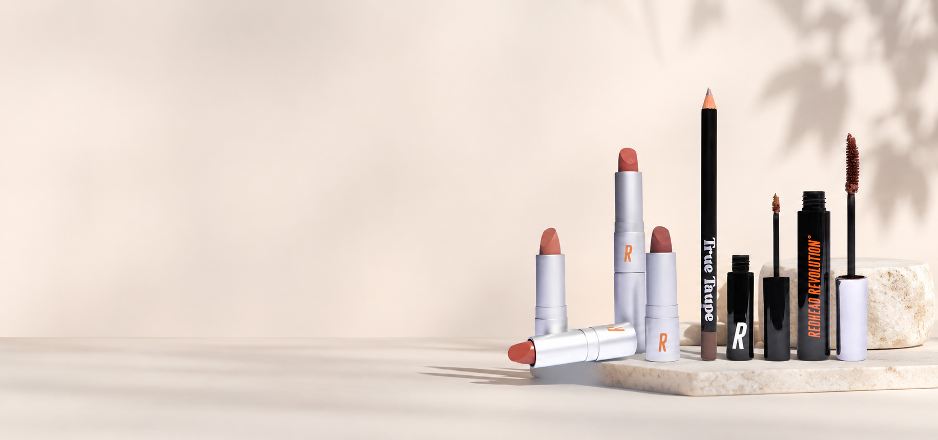 Redhead Revolution makeup essentials including lipstick, brow gel, mascara, and eyeliner arranged in a clean modern beauty display.slider_item_Tz6AzB